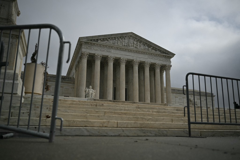 A low-angle view of the U.S. Supreme Court building, featuring its grand columns and marble steps, partially obscured by a metal barrier in the foreground.