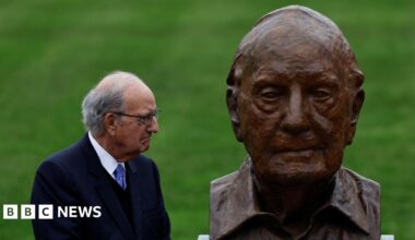 George Mitchell, with short grey hair, wearing glasses, a white shirt, purple tie and navy suit. He is looking to the right at a bust depicting himself.