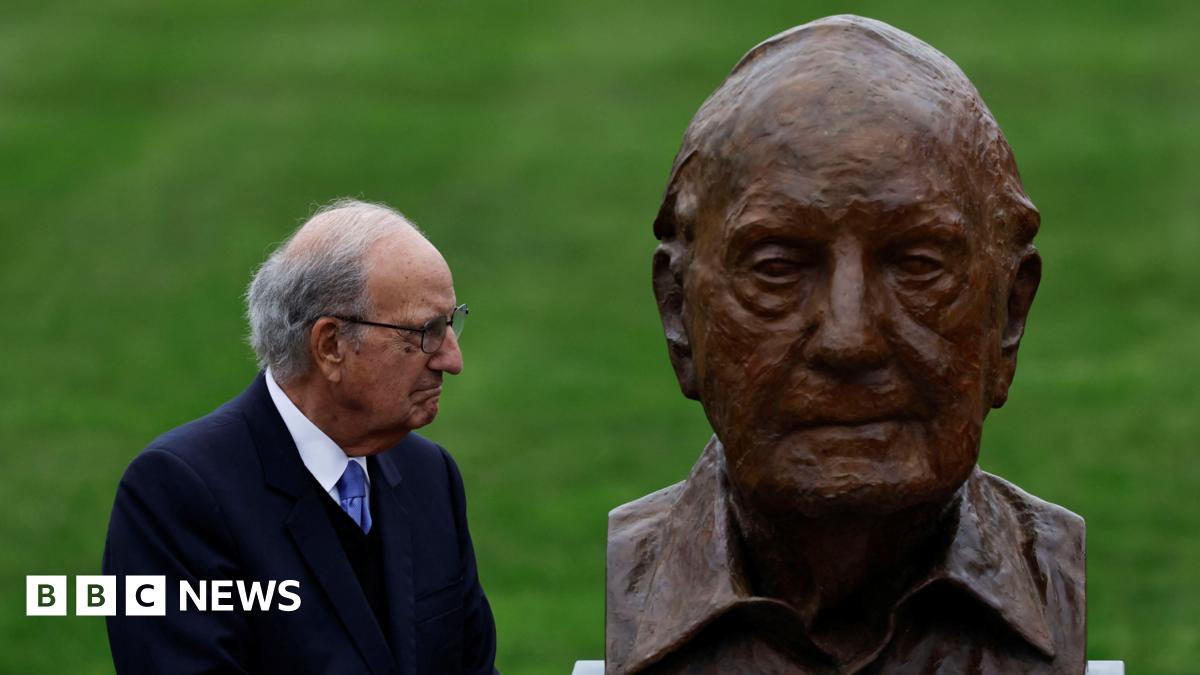 George Mitchell, with short grey hair, wearing glasses, a white shirt, purple tie and navy suit. He is looking to the right at a bust depicting himself.
