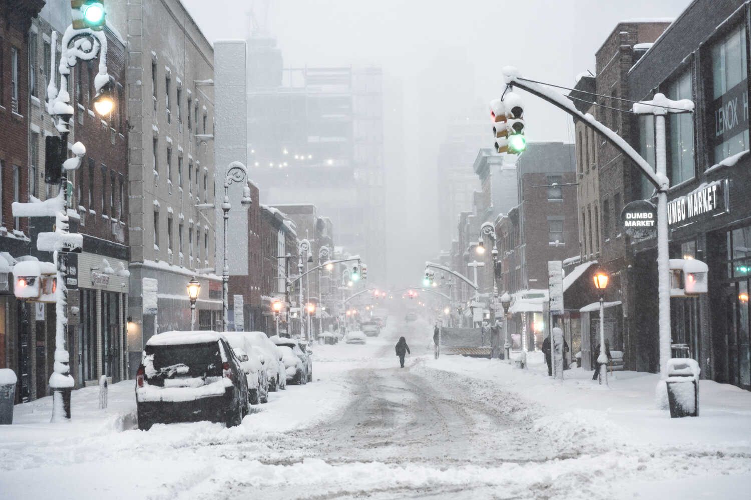 Snow falls on a street, covering vehicles and stop lights.