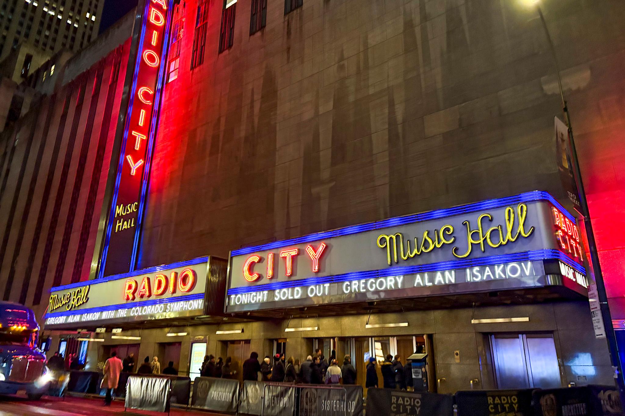 People line up along metal barricades outside. An awning above them reads "RADIO CITY MUSIC HALL" in neon lettering.