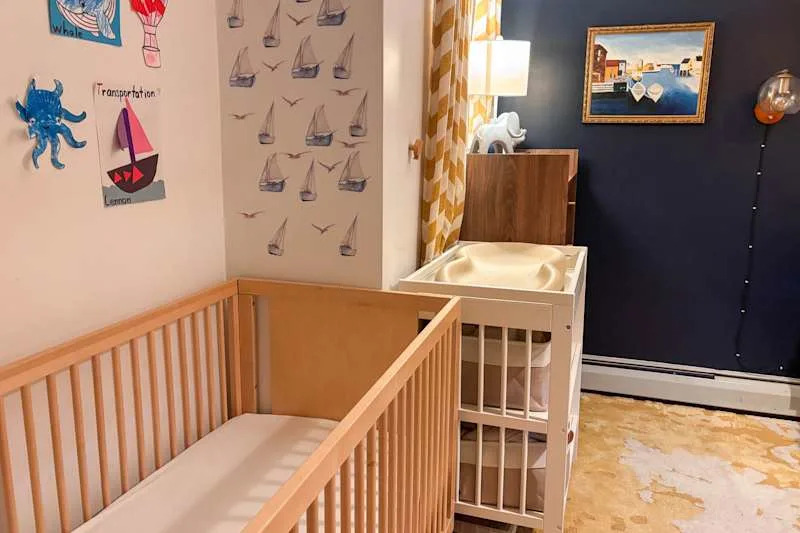 Nursery with a wooden crib, changing table, nautical wall decor, and a lamp beside a patterned curtain.