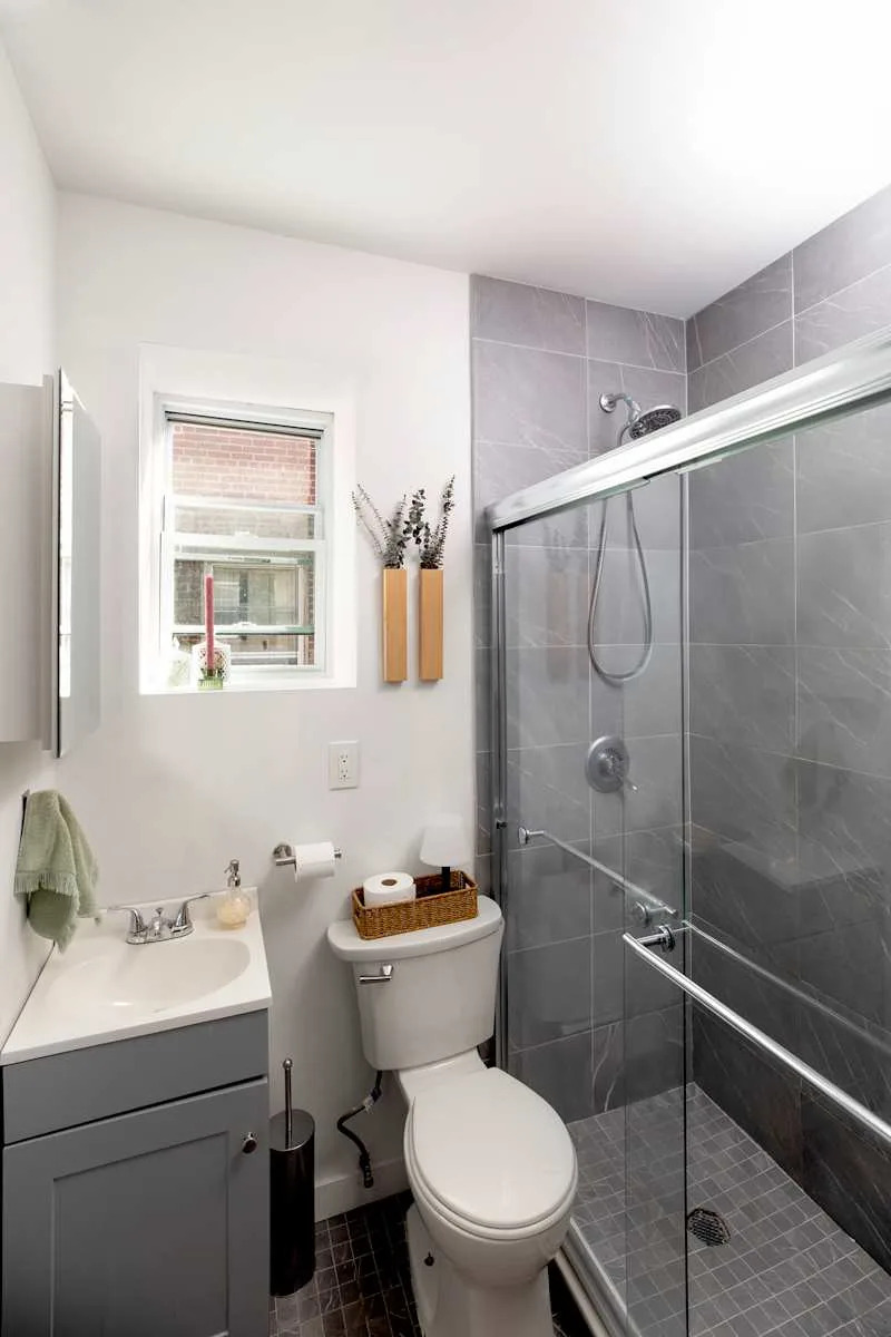 Modern bathroom featuring a glass shower, gray tiles, a white sink, and a small window with plants.