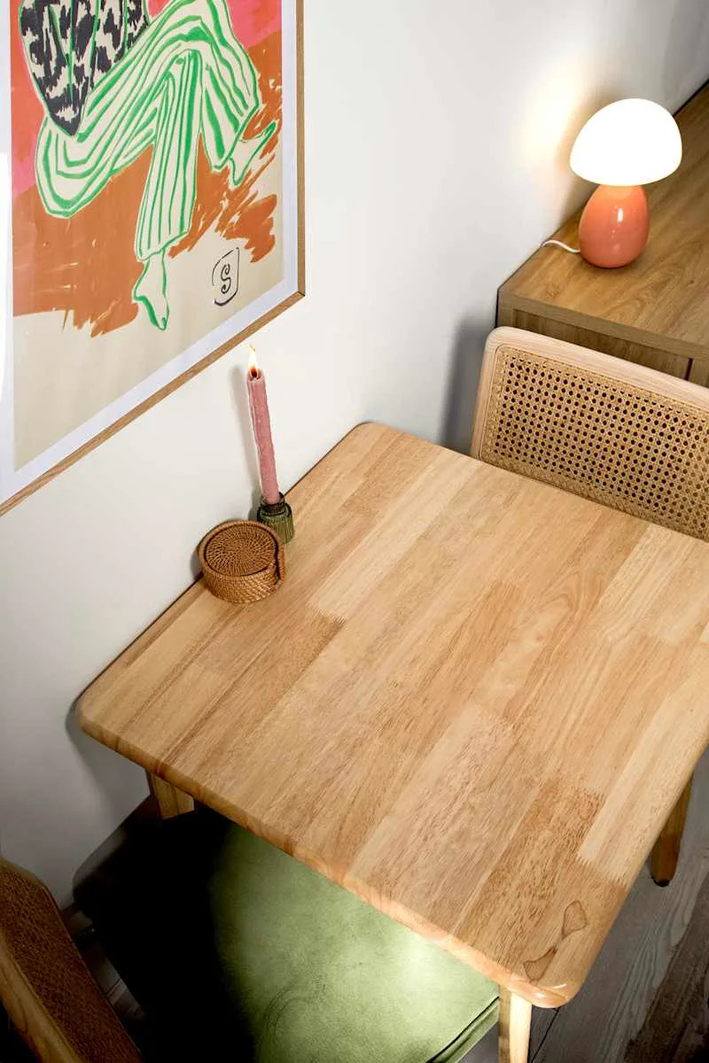 Wooden dining table with a pink candle, woven basket, green chair, and a colorful art piece on the wall.