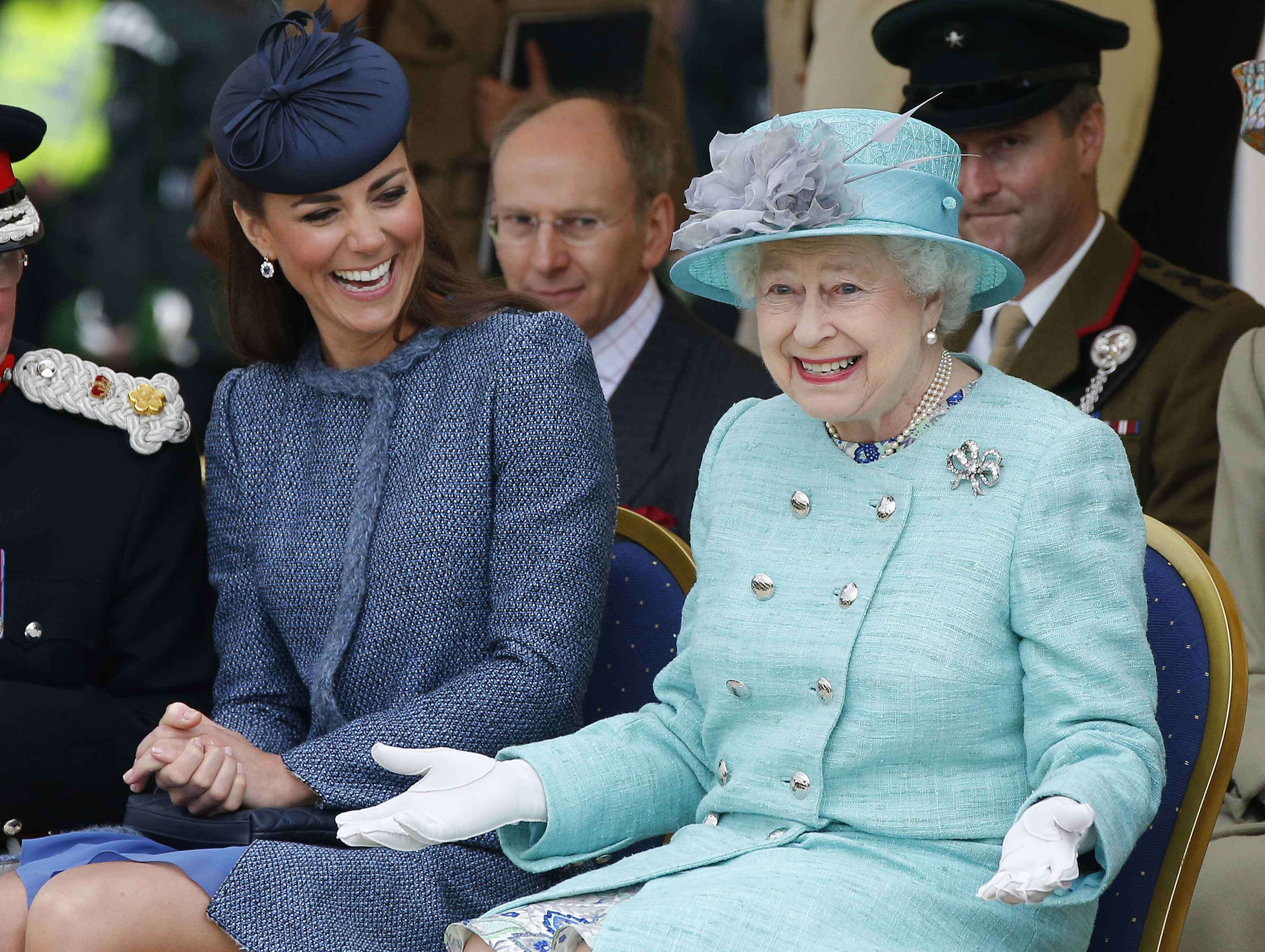 Kate Middleton wearing a blue coat sitting next to Queen Elizabeth, both are laughing