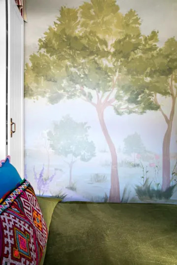 A cozy corner with a green cushion and colorful patterned pillow, featuring a mural of trees and flowers on the wall.