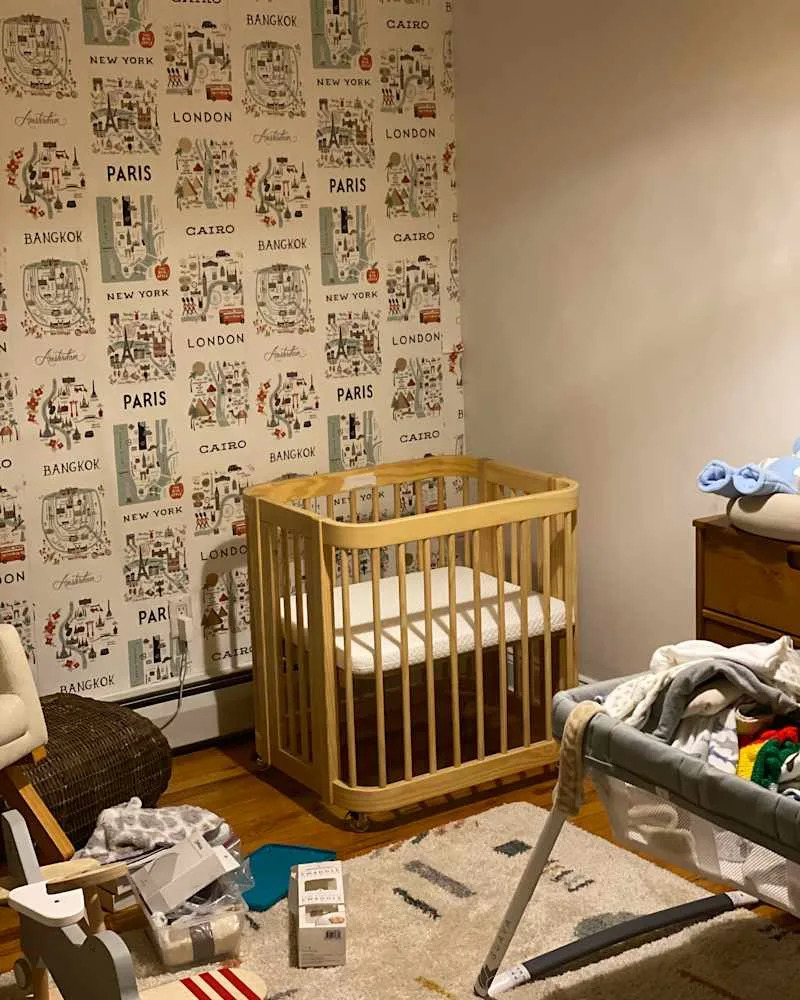 Nursery with a wooden crib, patterned wallpaper featuring city names, and scattered toys and blankets on the floor.