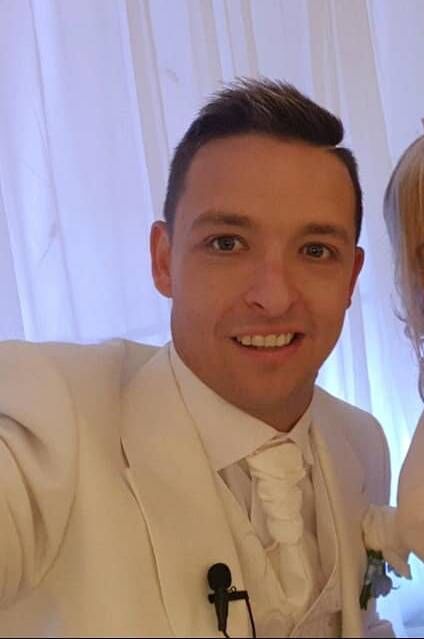 An individual dressed in formal attire, smiling brightly with a hint of a toothy grin, stands against a softly lit background.