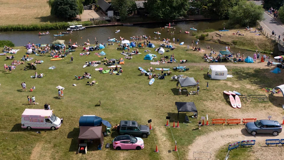 An aerial image taken of the River Stour, showing parked vehicles, an ice cream van, lots of people by river, tents, paddle boards, people in the river, and trees and grass areas. 