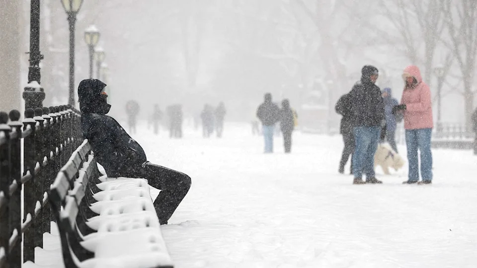 A bundled-up man rests on a park bench as snow falls steadily around him.