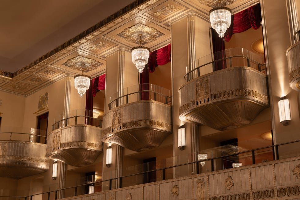 Interior view of a theater balcony with ornate decor and chandeliers.