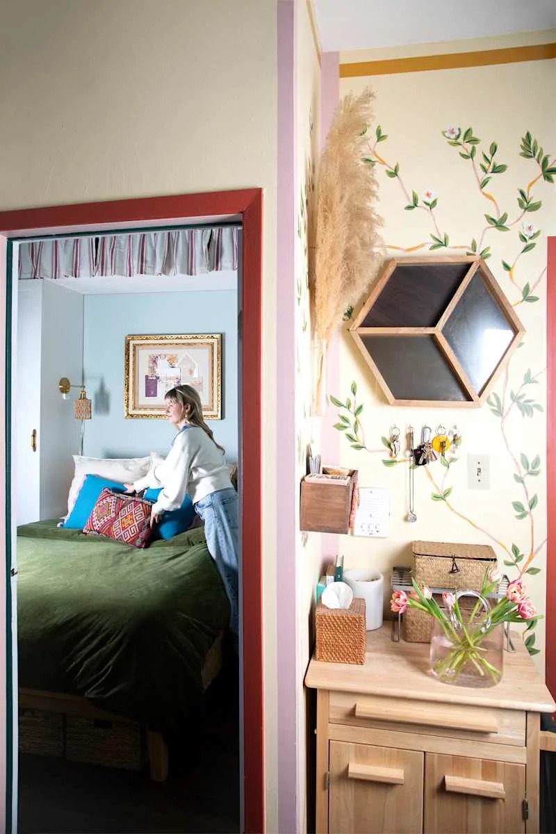 A woman arranging colorful pillows on a green bed in a cozy bedroom with floral wall decor and a wooden dresser.