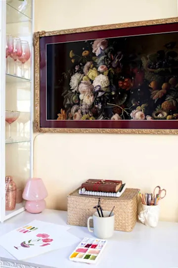 A workspace featuring a floral painting, pink lamp, sketch papers, watercolor palette, and decorative storage.