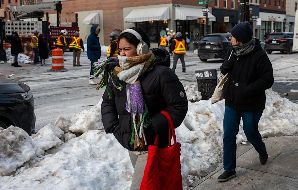 A woman is pictured screwing her face with a scarf tied around her mouth, and the hands in the pockets of her puffer jacket as she walks down a NYC street. Piles of shovelled snow are seen in the background
