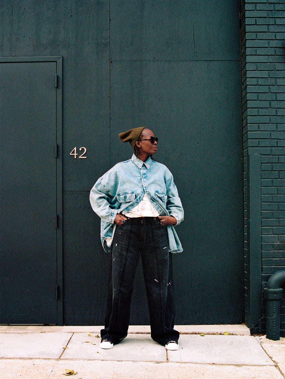 Person standing in front of a dark wall and door, wearing casual attire.