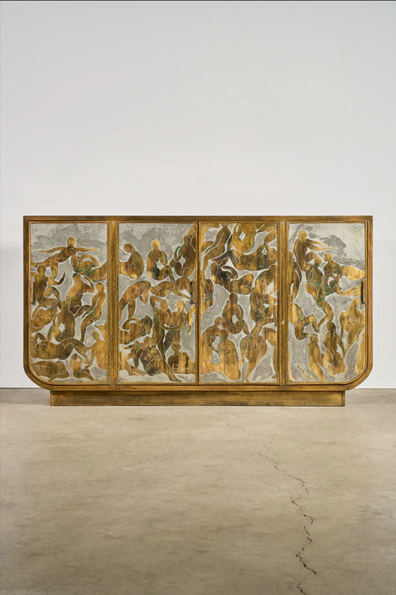 Vintage wooden cabinet with artistic, abstract carvings on the doors, set against a plain white wall and concrete floor.