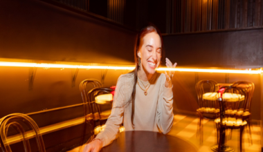 A young woman stands mid-street wearing a beige long-sleeve top, a black skirt, and black knee-high boots, with parked cars lining both sides.