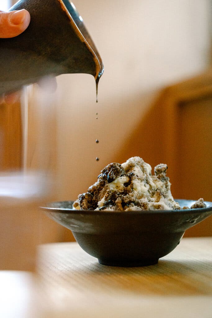 Dark drops of orgeat fall from a black pitcher into a black bowl of shave ice topped with corn pudding and a crumble of black sesame streusel.