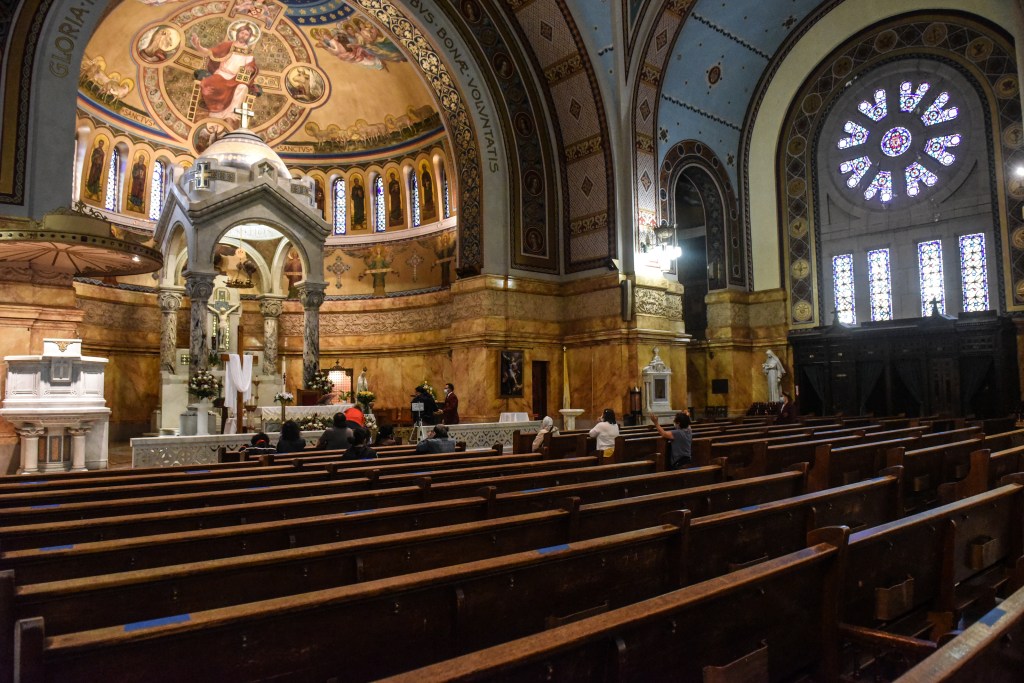 The interior of a church with ornately painted ceilings, stained-glass windows, and rows of wooden pews where a few people sit.