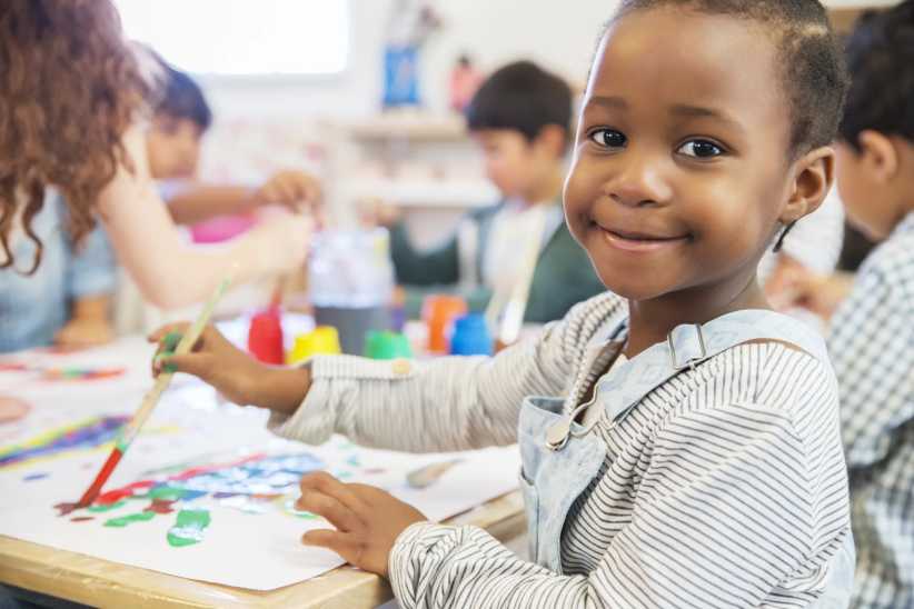 Pre-K student painting in class, looking at the camera and smiling