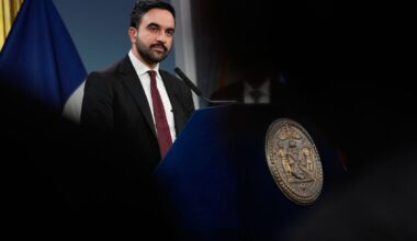 A photo of Zohran Mamdani, a light-skinned man with black hair and beard in a dark blue suit, maroon necktie and a white dress shirt, standing at the podium at a news conference with dark silhouettes framing the picture in the foreground.