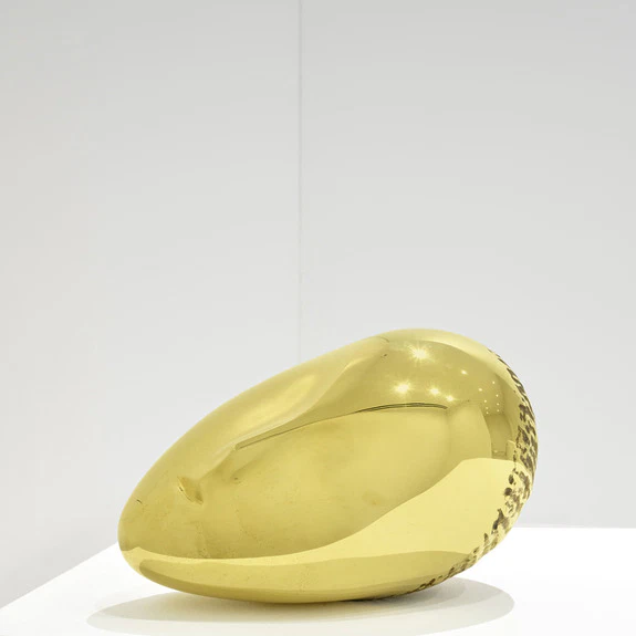Golden abstract sculpture displayed on a white platform against a minimalist background.