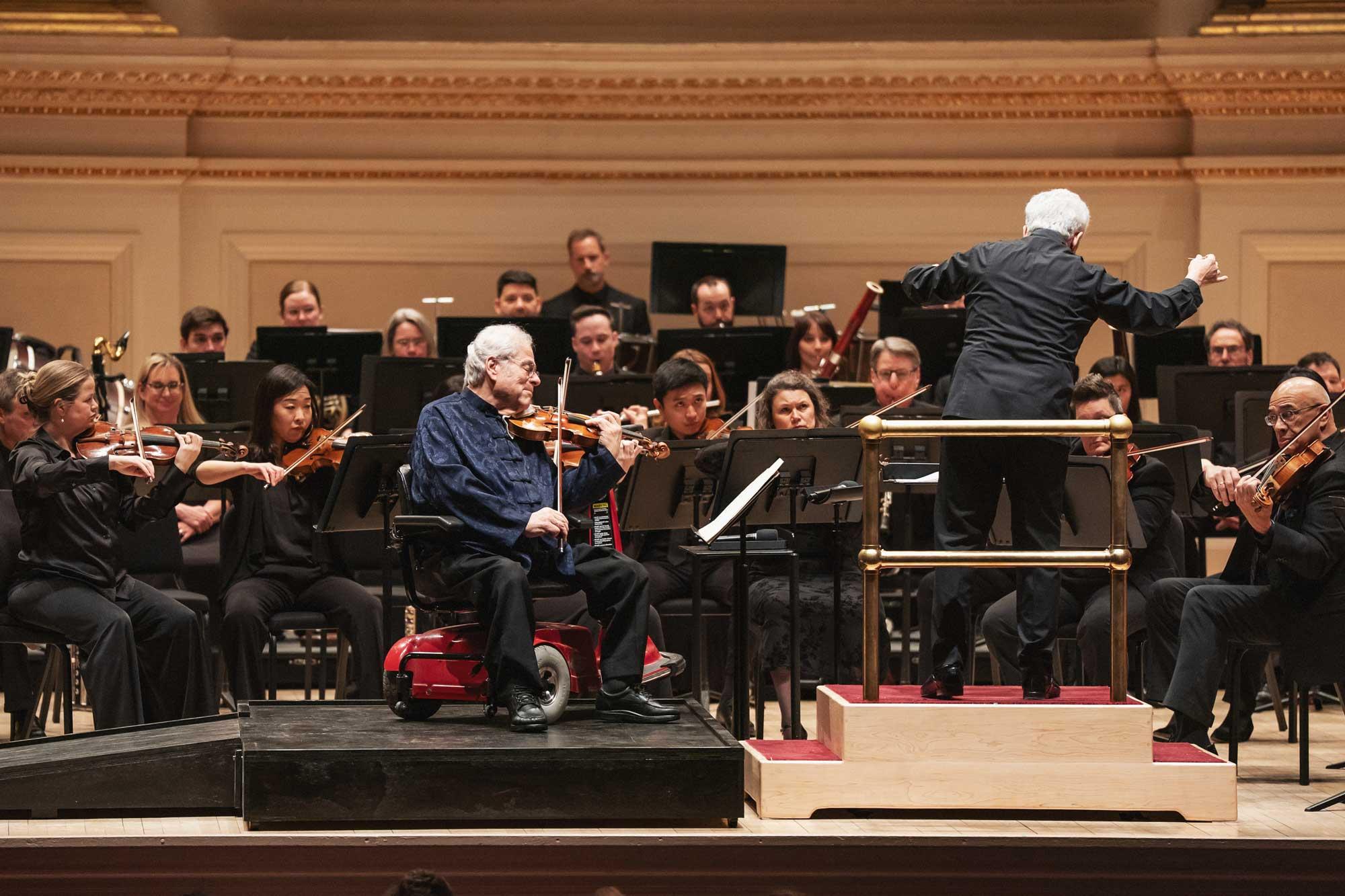 An older man sits on a red mobility aid and plays violin in front of an orchestra. Another older man faces toward the other musicians, conducting them.