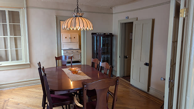 A dining room with a wooden table and six chairs, a hanging light fixture above, a glass-front cabinet, and a doorway leading to a separate room with white cabinetry at the Weeksville Heritage Center in Brooklyn.