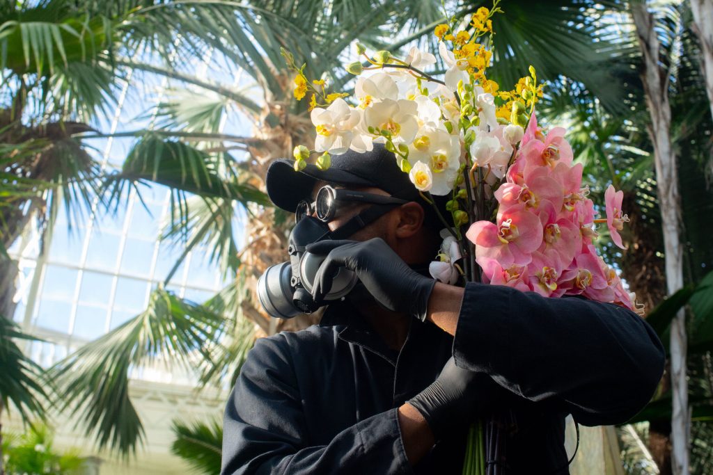 A mean wearing a black hat, goggles and respirator mask while holding pink and white orchids, looking to the left.