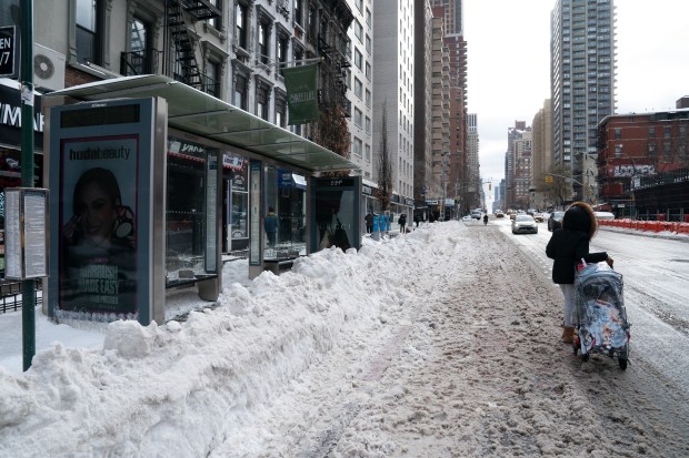 A pedestrian waits for a bus on the street by a bus stand inundated by snow on the corer of E. 87th St. and 1st Ave. in Manhattan on Monday, Jan. 26, 2026. (Barry Williams/ New York Daily News)