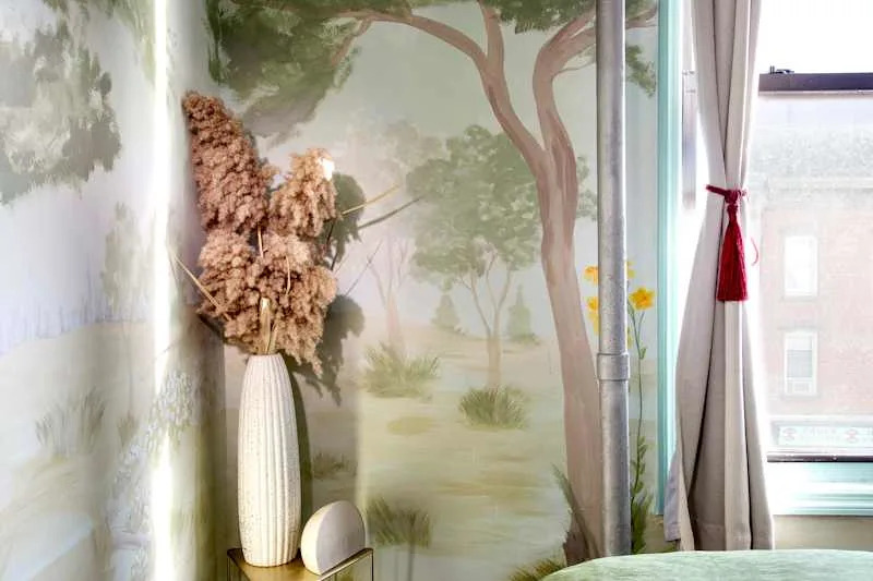 A corner of a room featuring a mural of trees, a tall white vase with dried flowers, and a small speaker on a gold table.