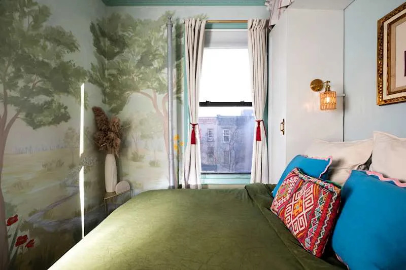 Cozy bedroom with green bedding, colorful pillows, a mural of trees, and a window with light curtains.