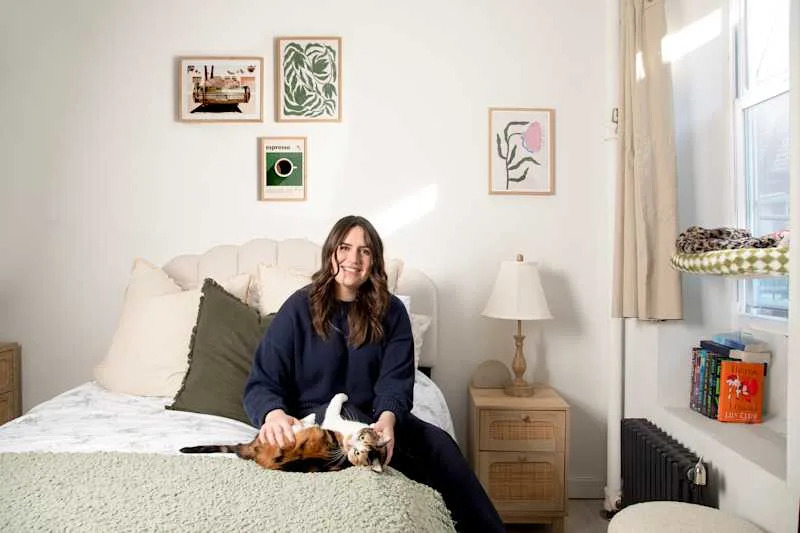 A woman sitting on a bed with a calico cat, surrounded by decorative pillows, framed art, and a lamp.