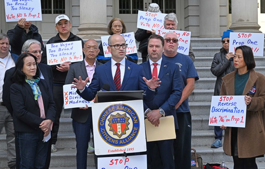 State Assemblyman Edward Ra speaks at a protest against Proposal 1, which aims to reduce ethnic imbalance in specialized high schools, surrounded by protestors holding signs that read "Vote NO to Prop 1" and "STOP Reverse Discrimination".