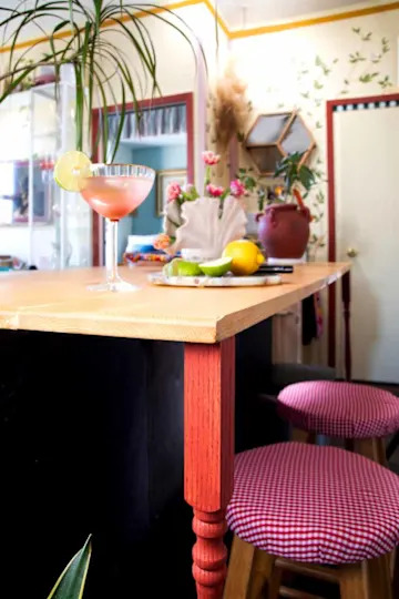 Cocktail glass with lime slice on a wooden bar, surrounded by fresh fruits and floral decor, with red checkered stools.