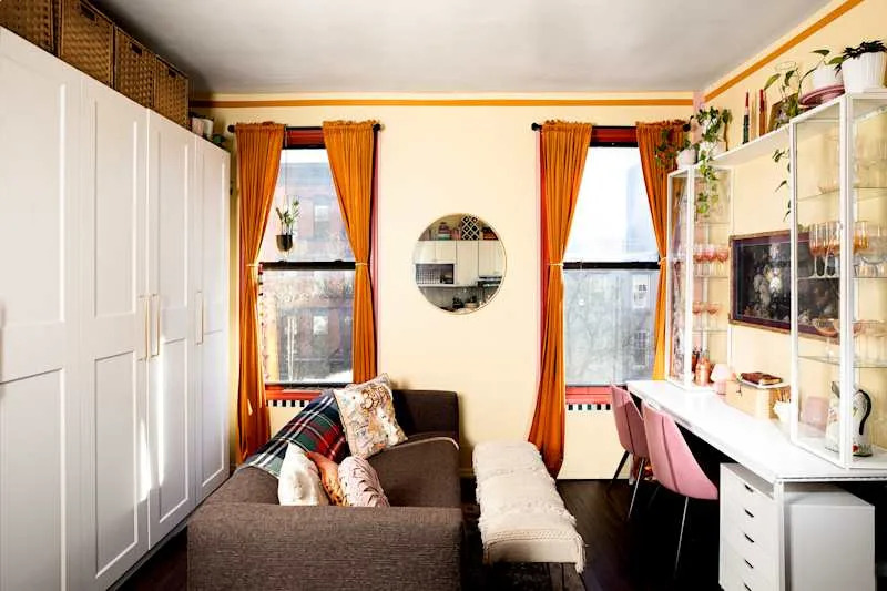 Cozy living space featuring a brown sofa with decorative pillows, orange curtains, a round mirror, and a white desk with pink chairs.