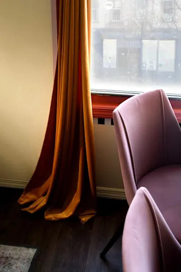 Warm orange curtain draping beside a pink upholstered chair near a window with a view of a street.