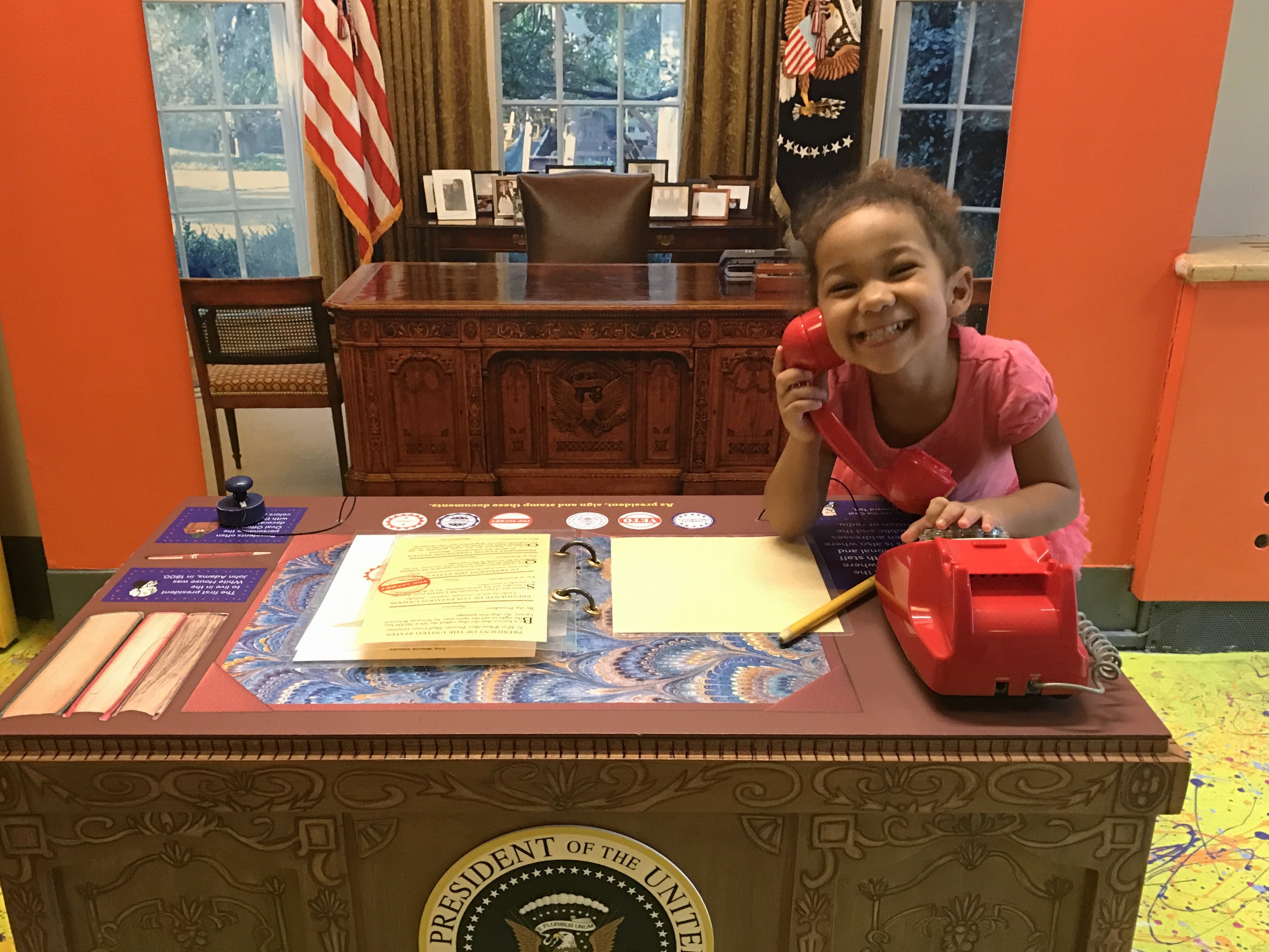 Girl in a pretend oval office behind a desk holding a red phone up to her ear with a big smile.