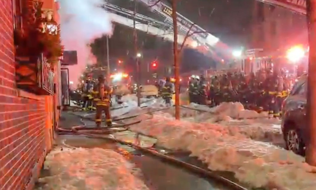 Nighttime photo of firefighters responding to a fire, with smoke visible from a building and fire hoses on the snowy ground.