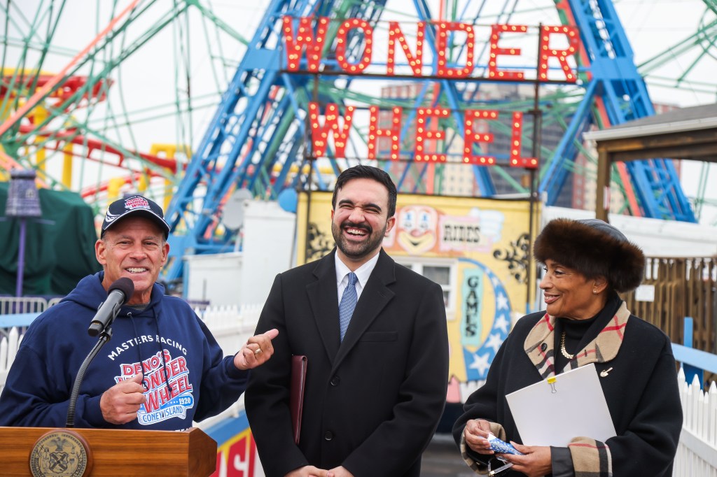 Dennis Vourderis speaks at a podium next to Zohran Mamdani and another woman, with the Wonder Wheel in the background.
