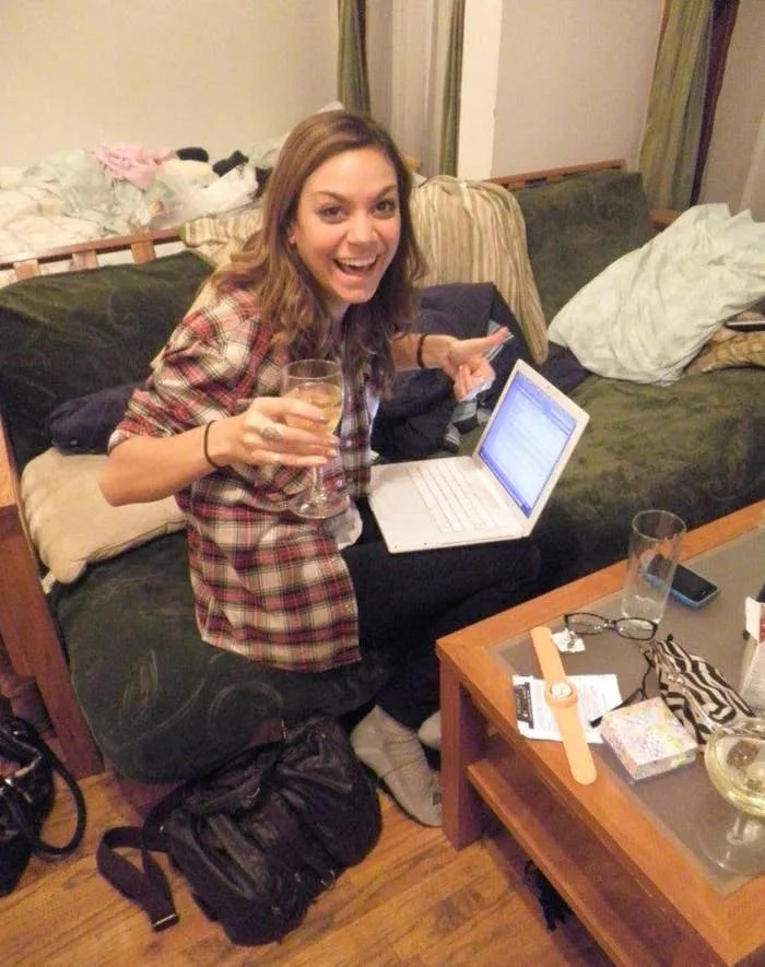 Person sitting on a couch, smiling with a drink in one hand and a laptop on their lap, surrounded by casual items on a coffee table