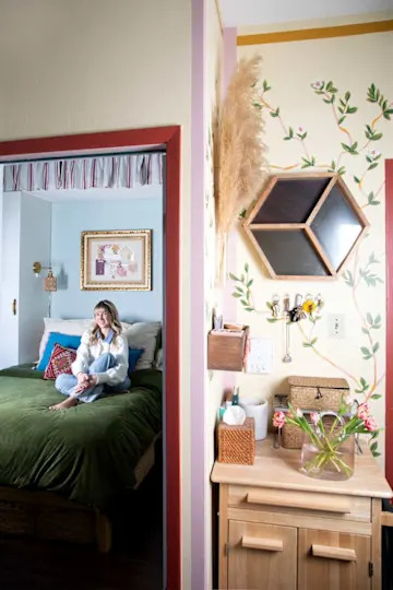 Cozy bedroom with a green bedspread, a woman sitting on the bed, and a decorative entryway with plants and storage.