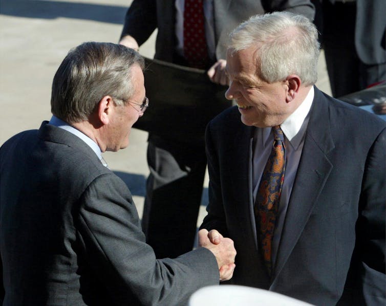 Two men, one white-haired, shake hands outdoors.