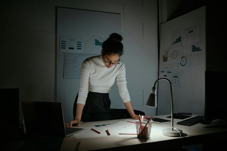 A woman standing at a desk in an office seemingly at night; the overhead lights are off, and she is only lit by a lamp