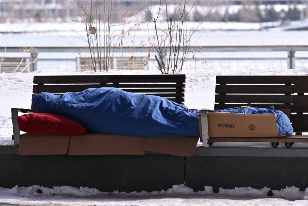 A homeless person sleeping on a park bench covered in a blue tarp with a red pillow, surrounded by snow.