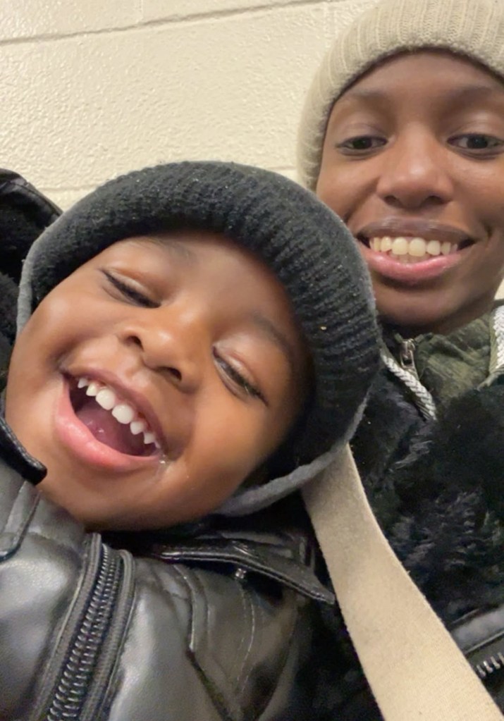 A young boy in a black knit cap smiling widely, with an adult woman in a beige knit cap smiling behind him.