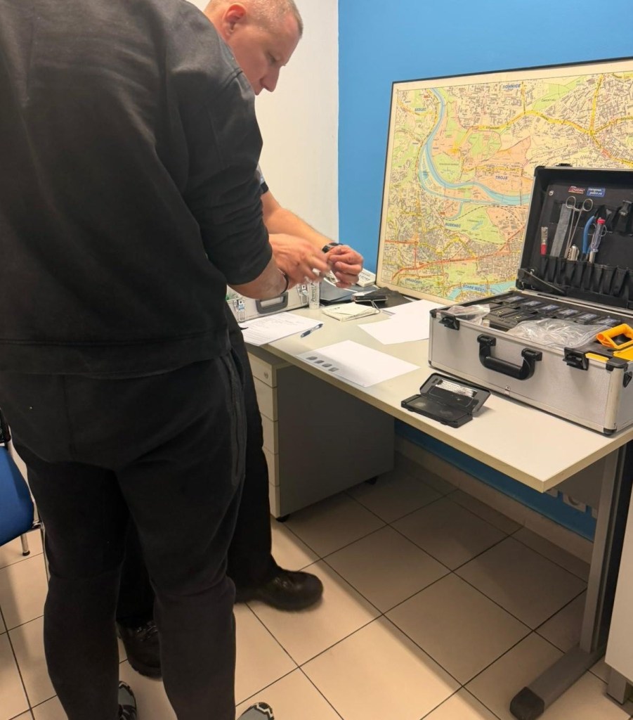 Two men standing at a desk, one is holding up his hands over a piece of paper, possibly for fingerprinting, next to a map and an open tool case.