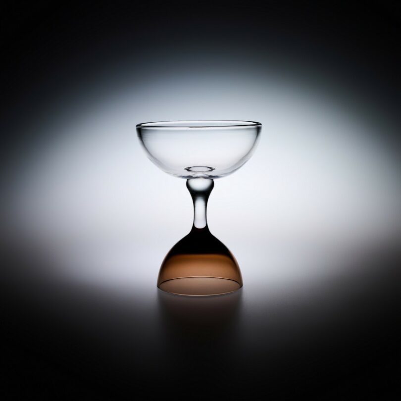 A clear glass coupe stands upright, with another identical coupe inverted and seamlessly joined at the base, against a softly lit gradient background