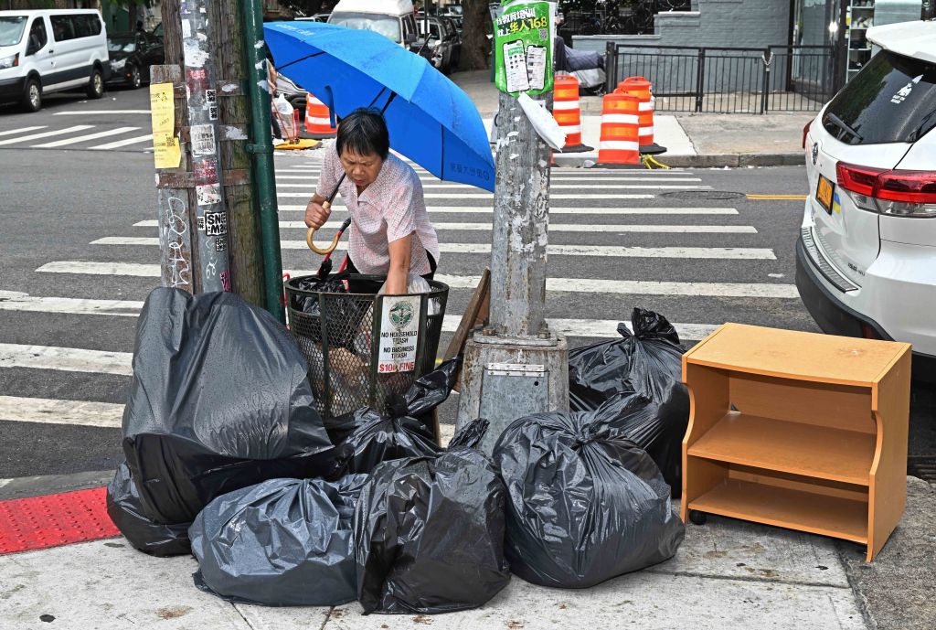 A woman with a blue umbrella places trash into a full wire mesh wastebasket surrounded by black garbage bags on a city sidewalk.