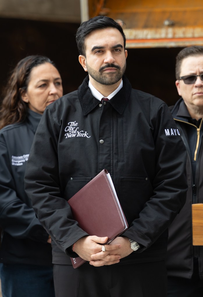 Zohran Mamdani, wearing a black jacket with "The City of New York" embroidered on it, holding a maroon folder at a press conference.